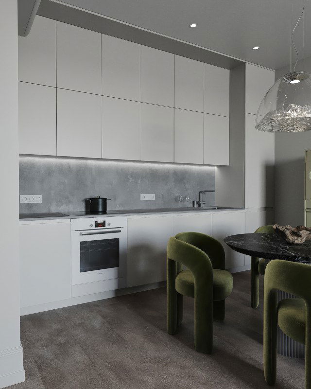 Вид на минималистичную белую кухню с встроенной техникой и серым фартуком, перед которой стоит тёмный круглый стол и зелёные бархатные стулья