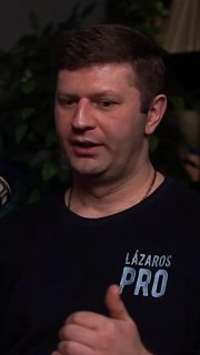 Крупный план мужчины в студии подкаста: тёмная футболка с надписью «LAZAROS PRO», жест рукой, фон с растениями и тёмной комнатой.