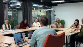 Группа слушателей и преподаватель за столом в офисной комнате на занятии по дизайну интерьера: ноутбук, блокноты и обсуждение проекта в практике курса.