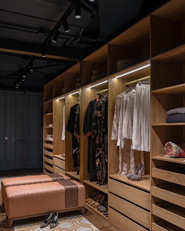 Ближний ракурс открытых секций гардеробной: одежда на вешалках, встроенная LED-подсветка, медовое дерево и пыльно‑синяя отделка коридора на заднем плане.