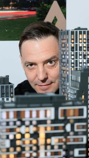 Крупный план лица дизайнера на фоне макета жилого комплекса «Архитектон», видны миниатюрные дома и фасады