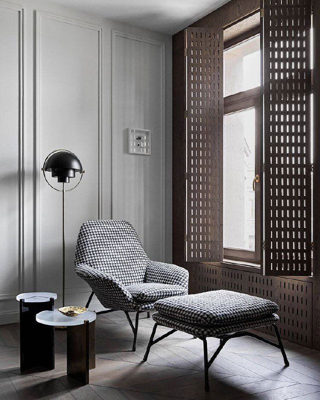 Сдержанный серый интерьер с перфорированными деревянными ставнями, креслом для отдыха и минималистичной приставной мебелью у окна.