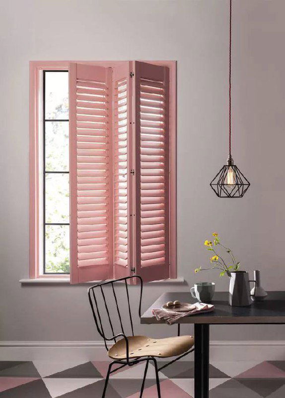 Минималистичная сцена: розовые ставни на окне, небольшой стол и подвесной светильник — аккуратное и нежное оформление маленького пространства.