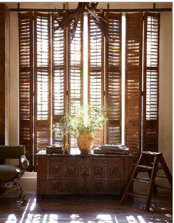 Тёплый интерьер с массивными темными деревянными ставнями, сквозь которые пробивается солнечный свет; перед окном декоративный сундук и зелень.