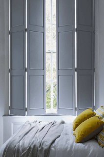 Спальная зона с высокими светло-серыми ставнями, мягкое одеяло и жёлтые декоративные подушки создают спокойный и современный образ окна.