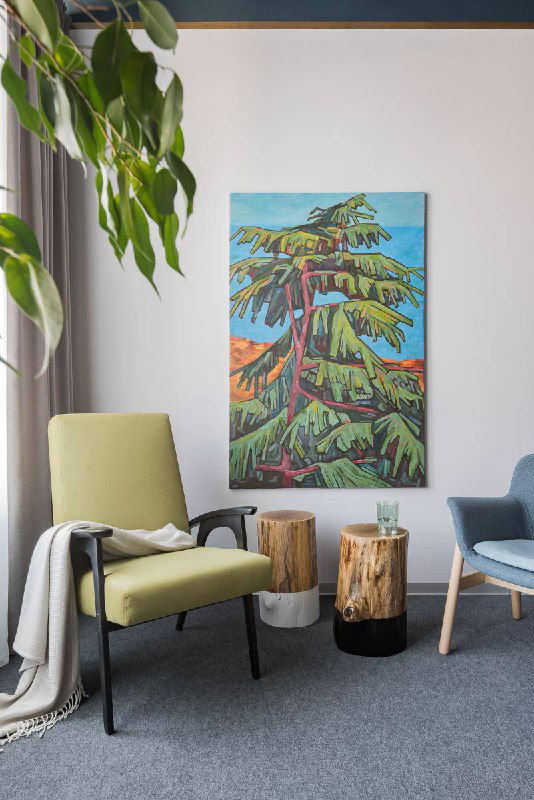 Уютный уголок с зелёным креслом и маленьким столиком, на стене висит крупная живописная картина с изображением дерева и яркими красками.