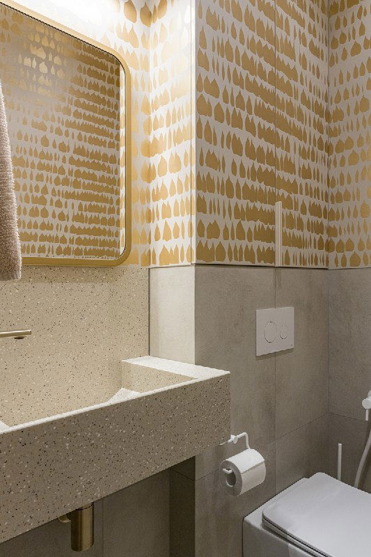 Угол гостевого санузла: золотой орнамент на стенах, панель инсталляции с кнопкой слива и держатель для туалетной бумаги на фоне фактурной столешницы.