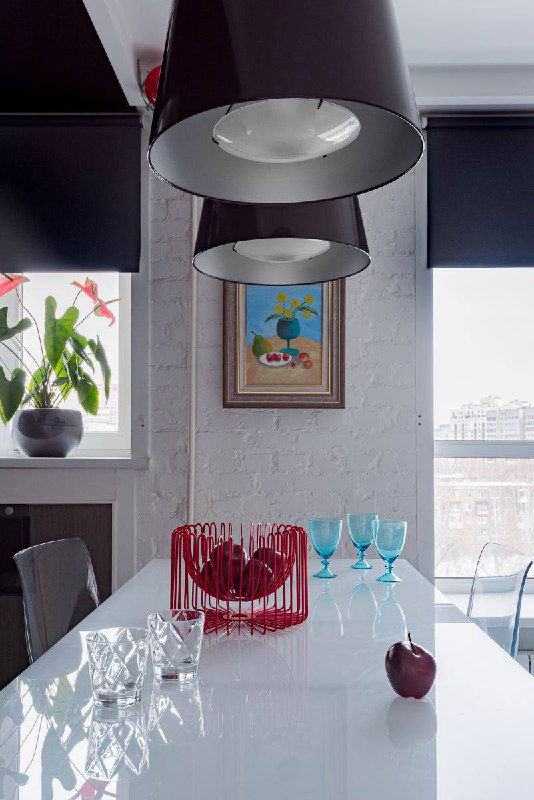 Современная столовая с белой глянцевой столешницей, подвесными светильниками и небольшой картиной в раме на стене, акцентный декор на фоне окна.