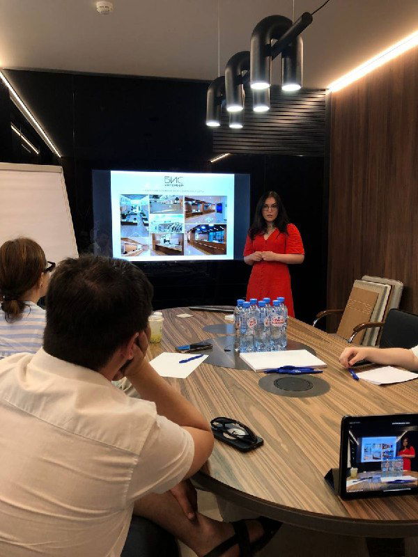 Презентация в переговорной: ведущая на фоне слайдов демонстрирует проекты, на столе вода и ноутбуки у слушателей, деловой формат встречи.