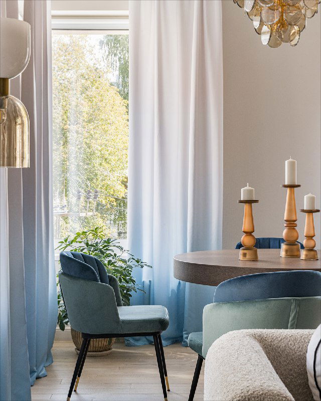 Угол кухни со синими креслами, растением у окна и овальным столом; светлые занавески и отражения придают глубину интерьеру.