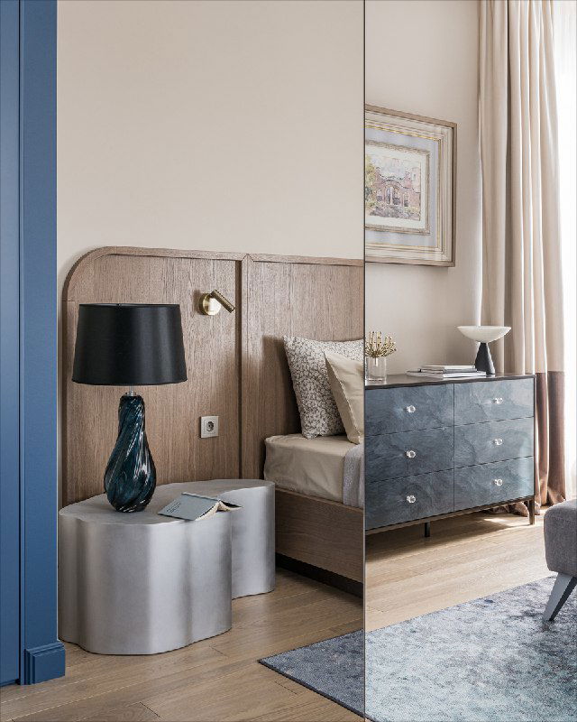 Спальня с зеркальными раздвижными дверями шкафа, отражающими прикроватную тумбочку и лампу, нейтральная палитра и текстуры.