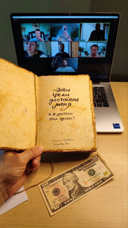 Рука держит старую тетрадь с вопросами и пометками; на заднем плане экран ноутбука с участниками и банкнота на столе.