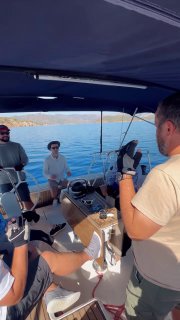 Несколько человек в кокпите парусной лодки во время регаты: рулевой и участники, вид на спокойную воду, части такелажа и береговой горизонт на заднем плане.