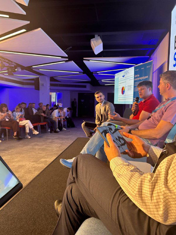 Панельная дискуссия в зале: несколько спикеров на сцене, экран с слайдами и участники в зале, вид с места в аудитории.