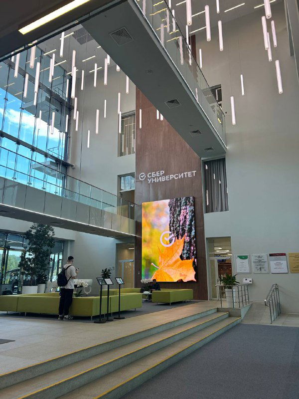 Интерьер вестибюля СберУниверситета: просторное фойе с лестницей, большой вертикальный LED-экран и современная мебель в нейтральных тонах.