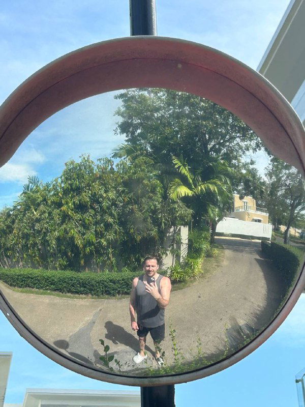 Отражение в выпуклом зеркале: автор и окружающая дорога с растительностью, визуальный акцент на самонаблюдении и паузе в пути.