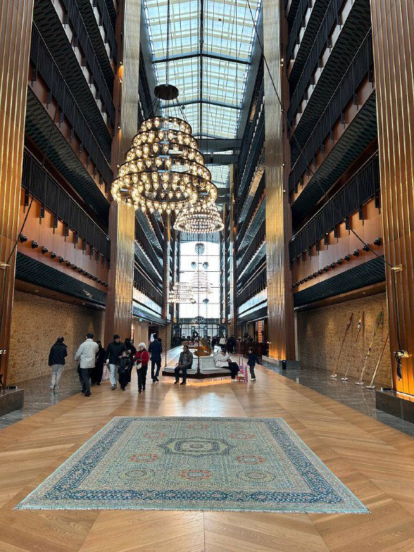 Просторный интерьер с большой хрустальной люстрой и ковром в центре — возможно лобби отеля или торгового центра в Стамбуле, много людей на фоне.