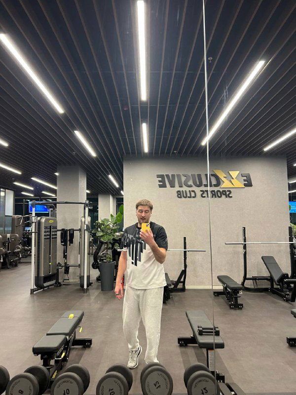 Тренажёрный зал с зеркалами, спортивным оборудованием и человеком в спортивной одежде, отражающимся в зеркале — кадр из спортклуба в поездке.