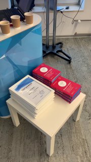 Столик с раздаточными материалами Школы трекеров: стопки книг и сертификатов, рядом несколько бумажных стаканчиков в офисной обстановке.