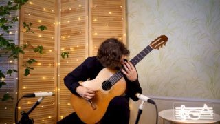 Гитарист в уютной комнате играет на классической гитаре; рядом два микрофона и тёплая декоративная подсветка — атмосфера репетиции и записи.