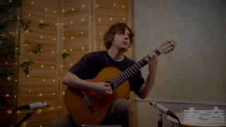 Музыкант сидит с классической гитарой в уютной студии, исполняет «Романс»; на фоне видны микрофон и декоративная гирлянда.