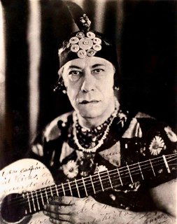 Чёрно‑белая винтажная фотография человека в сценическом костюме с гитарой: крупный план лица и дека инструмента, театральный образ.