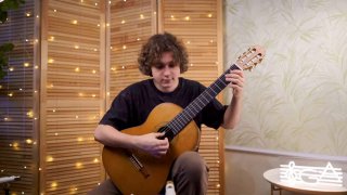 Гитарист в домашней студии играет классическую гитару, на фоне — гирляндные огни и декоративный музыкальный знак на столе.