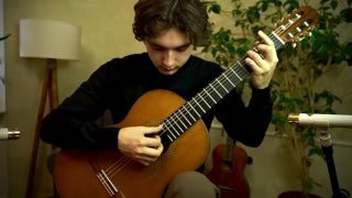 Исполнитель играет на классической гитаре в комнате: крупный план левой и правой руки на грифе и корпусе инструмента, камерная спокойная обстановка.