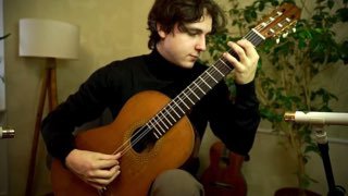 Исполнитель играет на классической гитаре в уютной домашней обстановке: видны корпус и гриф гитары, струны, микрофон и лампа.