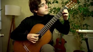 Гитарист в комнате репетиций играет классическую гитару вблизи микрофона; на заднем фоне видны ещё два инструмента и растения