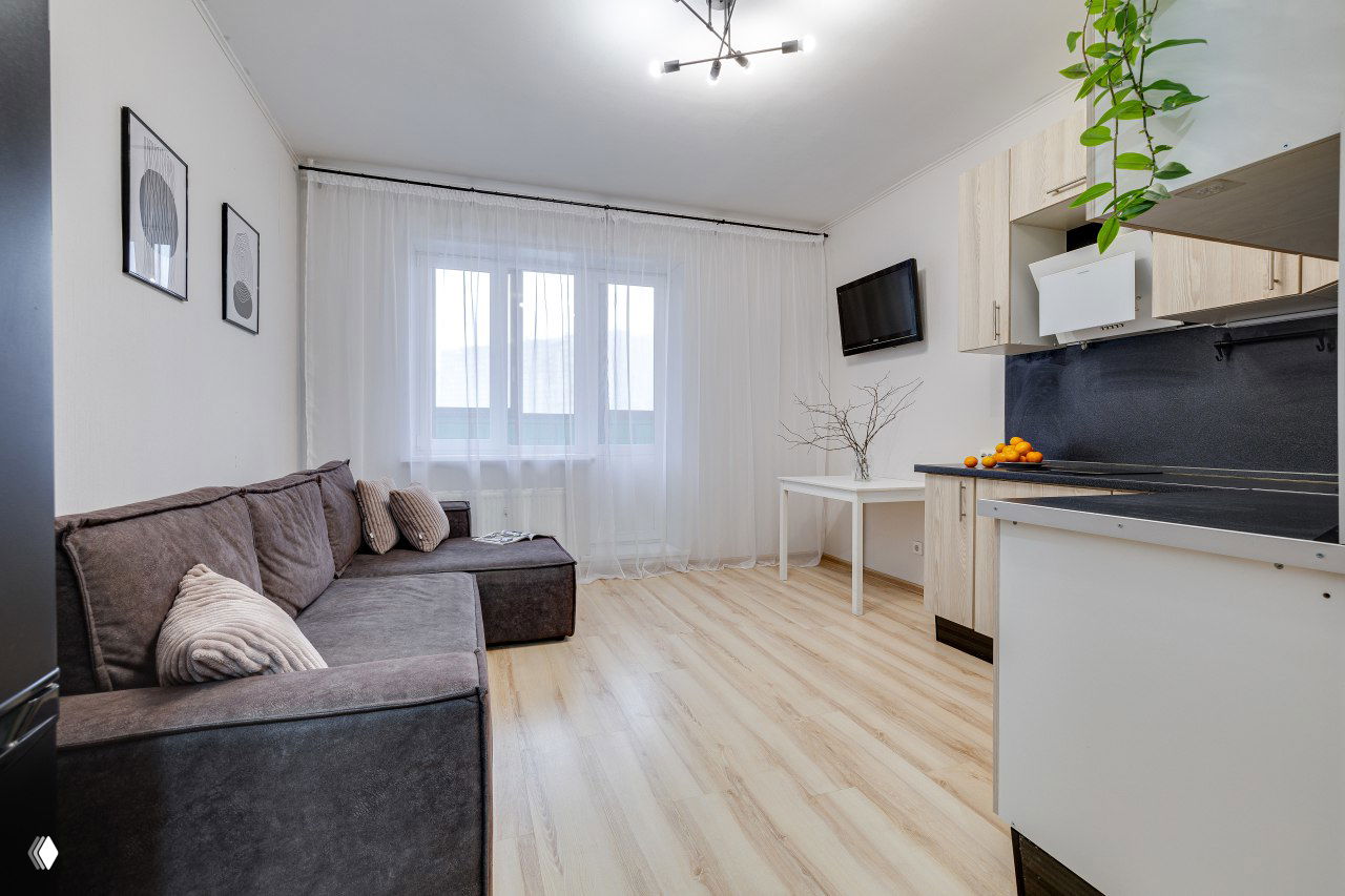 Широкий план гостиной-кухни: окно с тюлем, диван, небольшой стол и кухонный гарнитур — светлое, аккуратное помещение, оформленное для показа.