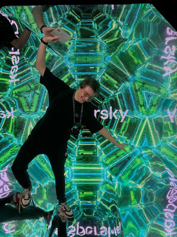 Посетитель позирует внутри инсталляции на стенде OffZone 2024, яркие зелёные геометрические узоры и зеркальные эффекты вокруг.