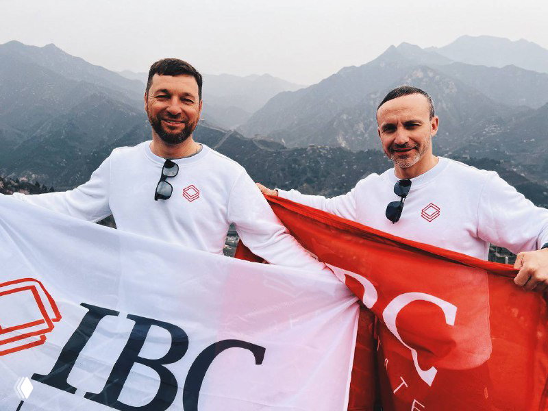 Два представителя IBC Real Estate держат фирменные флаги на фоне горного пейзажа во время деловой поездки в Китай; корпоративная фотография на открытом воздухе.