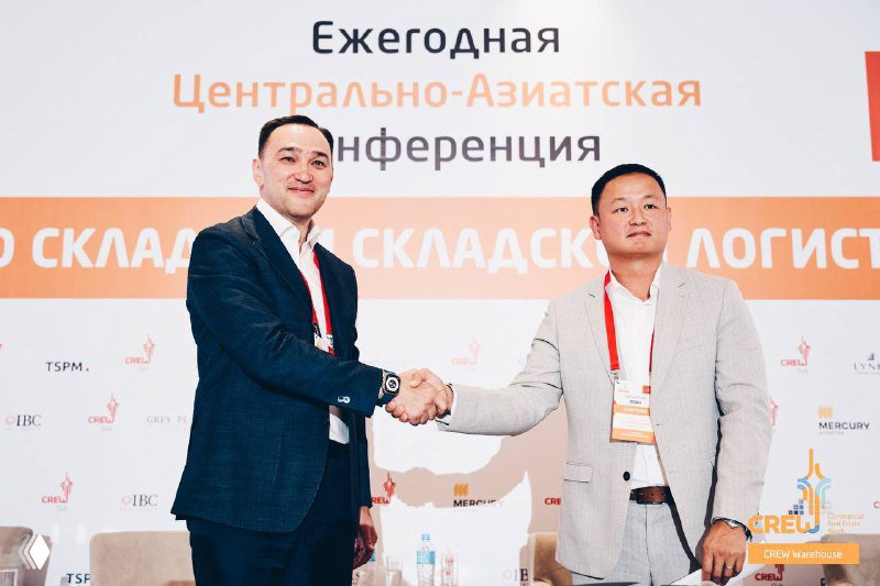 Два человека пожимают руки на сцене конференции CREW Warehouse на фоне баннера «Центрально-Азиатская конференция», деловая атмосфера и подписание сделки.