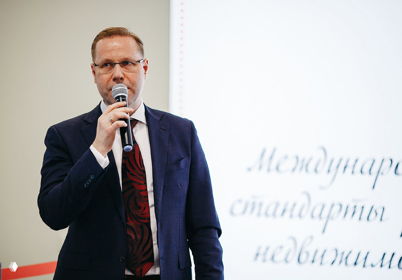 Дмитрий Шелковский держит микрофон и выступает на деловой конференции по недвижимости в Москве; мужчина в костюме на фоне презентации.