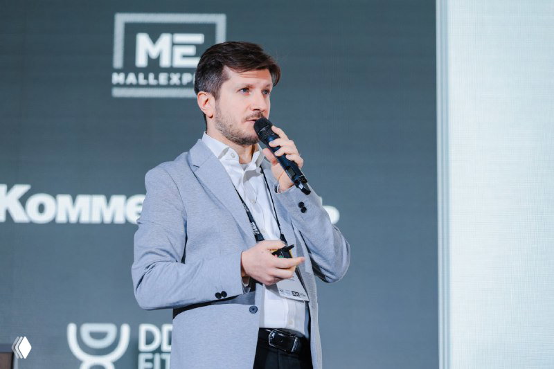 Докладчик на сцене MALLPIC 5.0 в светлом пиджаке держит микрофон и презентует слайд, кадр с мероприятия в формате крупного плана.