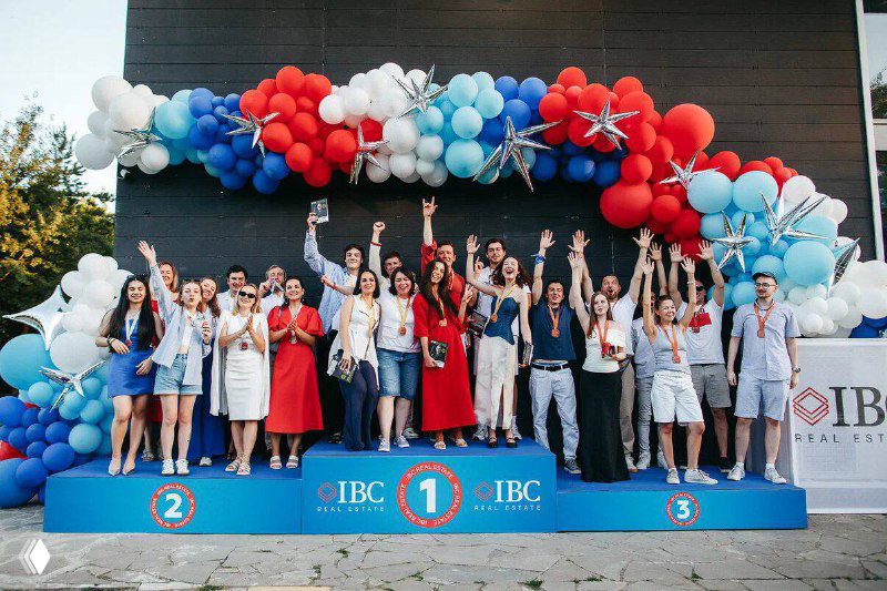 Подиум с победителями и участниками церемонии награждения: команды поднимают руки, фон украшен логотипом IBC и праздничными шарами.
