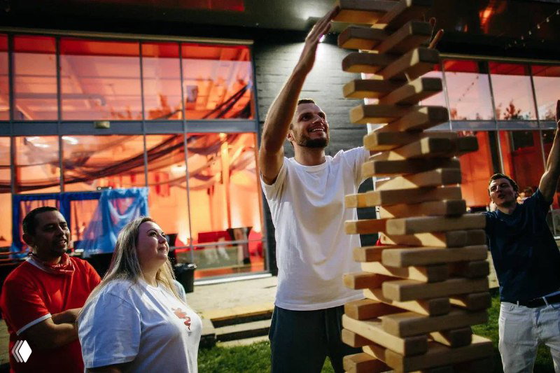 Развлекательная зона вечера: крупная настольная игра Jenga и сотрудники, улыбающиеся и участвующие в командных активностях после спортивной части.