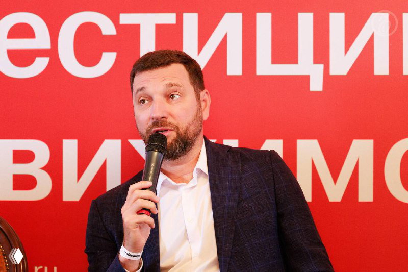 Крупный план Алексея Ефимова с микрофоном на фоне красного баннера «Инвестиции в недвижимость» во время выступления на мероприятии.