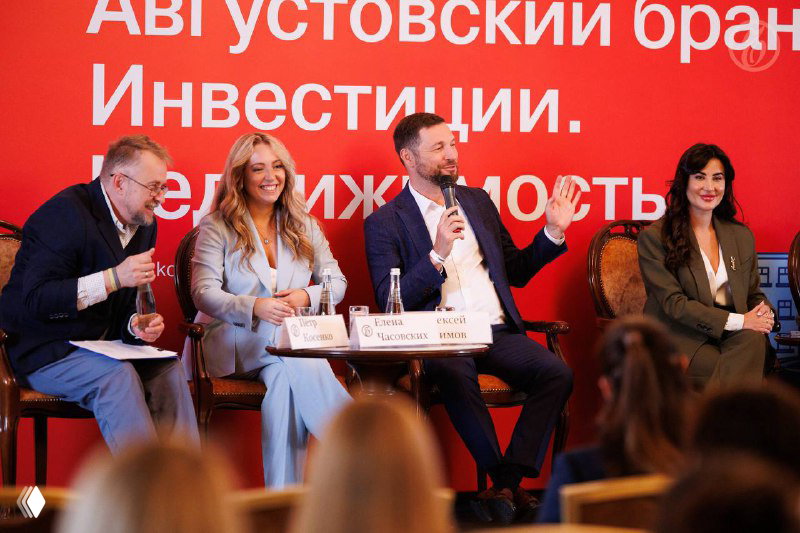 Три участника дискуссии на сцене ведут обсуждение: жесты, улыбки и живой диалог на фоне брендированного баннера мероприятия.
