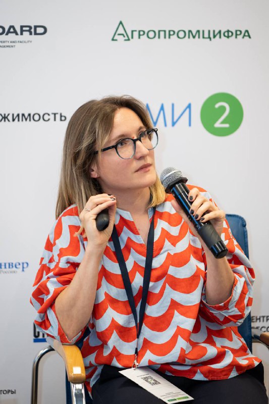 Крупный план докладчицы с микрофоном в руке на фоне бренд-волла; докладчица объясняет тезисы выступления и показывает внимание аудитории
