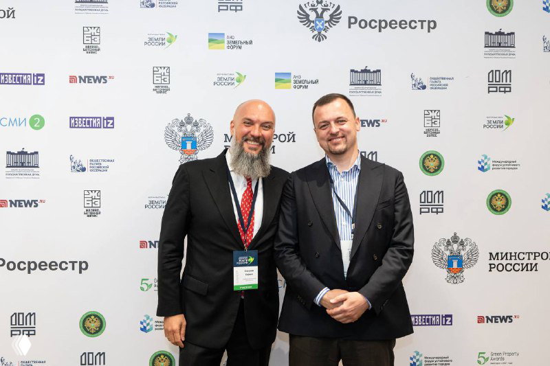 Два участника у бренд-волла форума улыбаются на фото на фоне логотипов партнёров и организаторов мероприятия, официальный фон мероприятия