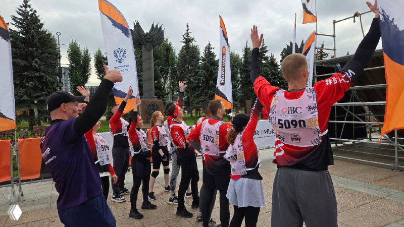 Сцена празднования команды IBC у сцены и флагов мероприятия: участники ликуют, поднимают руки и обмениваются эмоциями после этапа гонки.