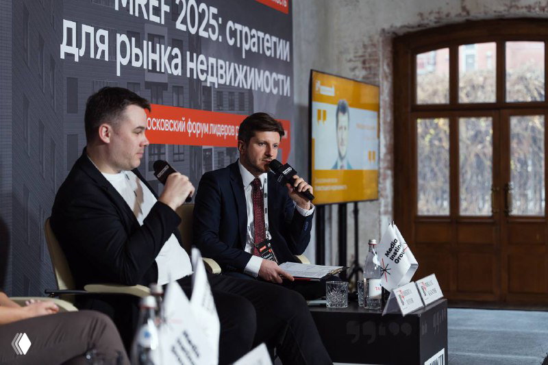 Два спикера ведут разговор в сессии «Инвестиции в недвижимость» на XIX Московском форуме: ведущий и приглашённый с микрофонами, на заднем плане экран и брендирование.