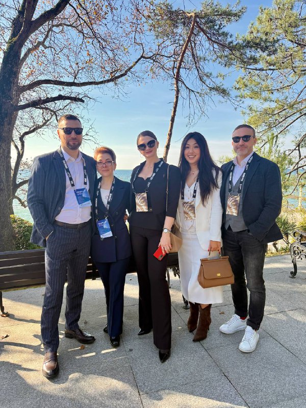 Командное фото IBC Real Estate на прогулке в Сочи во время MALLPIC SOCHI 6.0: участники в деловой одежде на фоне морского пейзажа и деревьев.