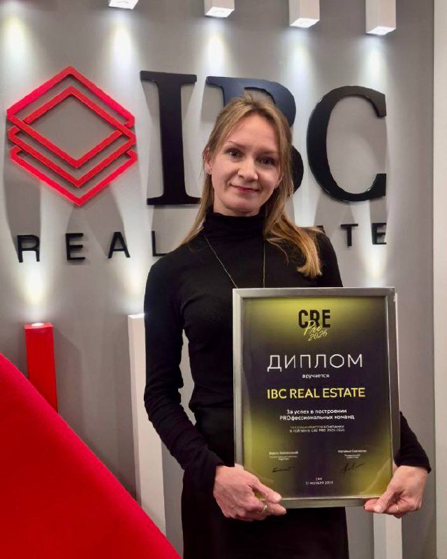 Женщина держит диплом CRE PRO 2026 на фоне логотипа IBC Real Estate; фотография демонстрирует награду и корпоративный интерьер, подчёркивая профессиональное признание компании.