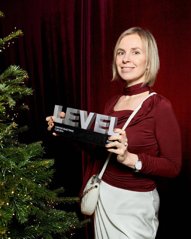Елена Шохина держит награду с надписью «LEVEL» на фоне бордовой занавеси и украшенной рождественской ёлки во время церемонии Level Group.