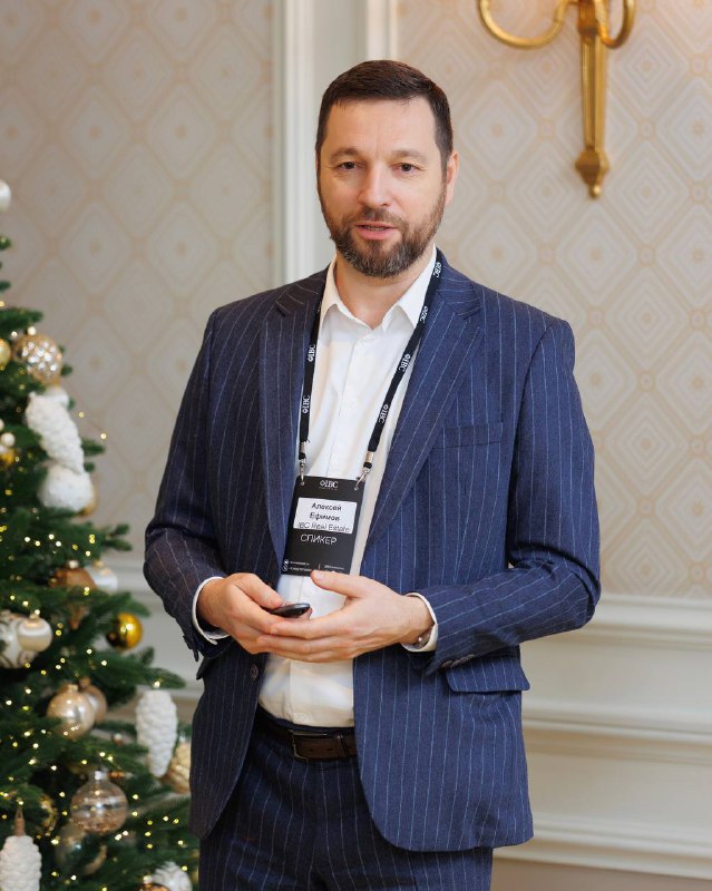 Крупный план выступающего спикера в костюме на пресс‑бранче IBC Real Estate; на заднем плане новогодняя ёлка и интерьер зала отеля.