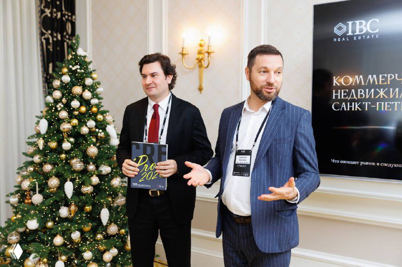 Два спикера на сцене у брендовой панели IBC Real Estate проводят диалог перед аудиторией в элегантном зале отеля.