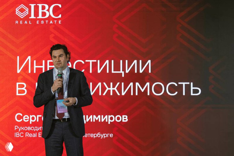 Сергей Владимиров на фоне красной сценической панели с логотипом IBC Real Estate и надписью «Инвестиции в недвижимость» во время доклада